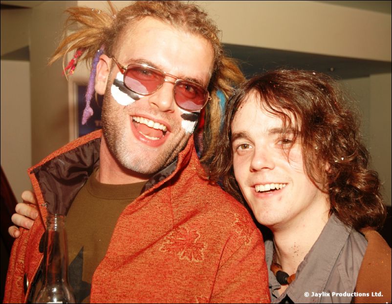 A smiling man with face paint, tinted glasses and dreadlocks stands beside a man with shoulder-length hair
