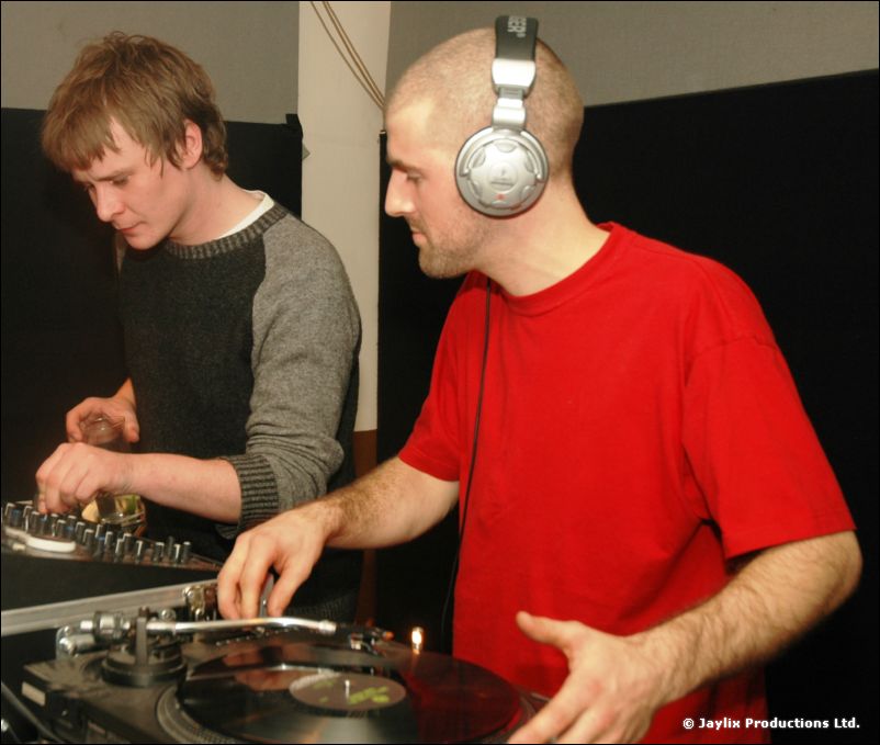 Two DJs at turntables, one in a red t-shirt wearing headphones, the other in a grey jumper