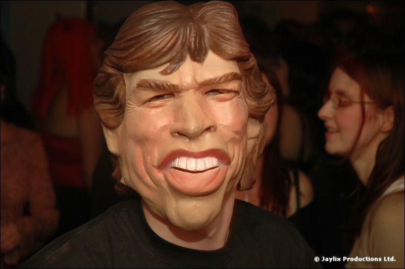 A person wearing a rubber mask of a face with shaggy hair and a wide grin