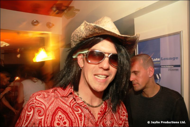 A person in a cowboy hat, long black wig, sunglasses and red patterned shirt smiles at the camera