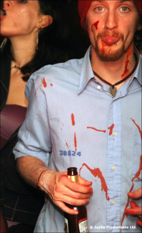 A man in a blood-spattered blue shirt with blood on his face sticks out his tongue, holding a bottle