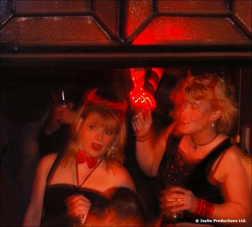 Two women wearing devil horns, one with a red bow tie, in a dimly lit red space
