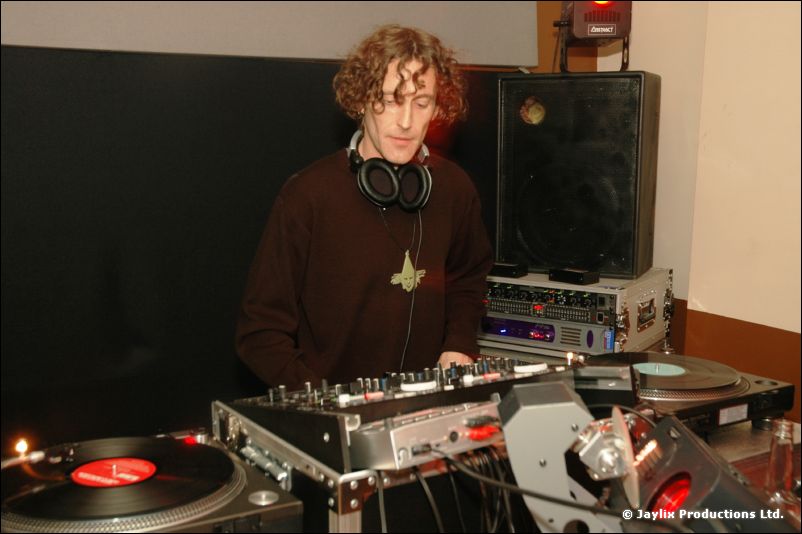 A DJ with curly hair and headphones at turntables against a black wall