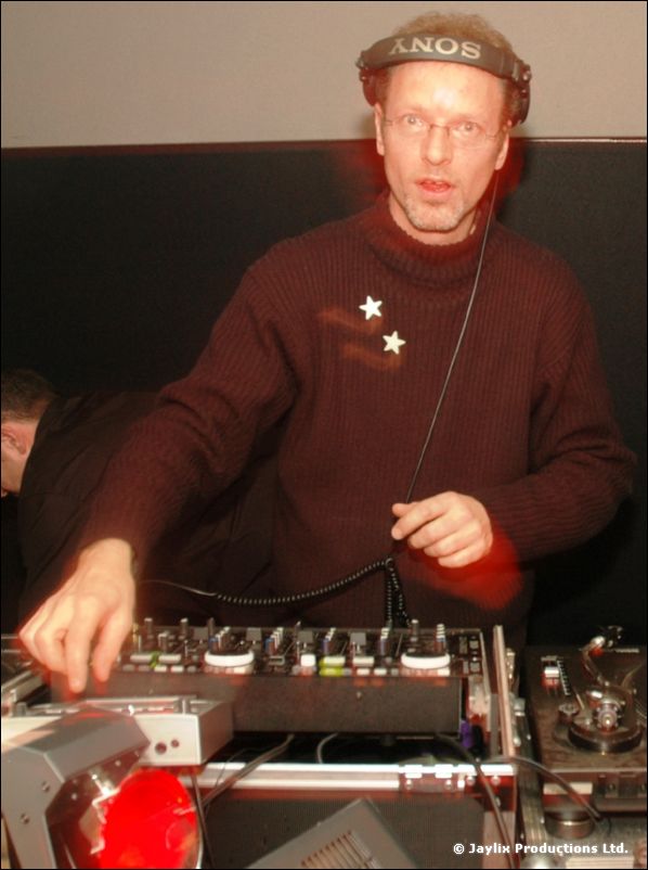 A DJ with glasses and headphones in a burgundy jumper with two stars, working a mixer