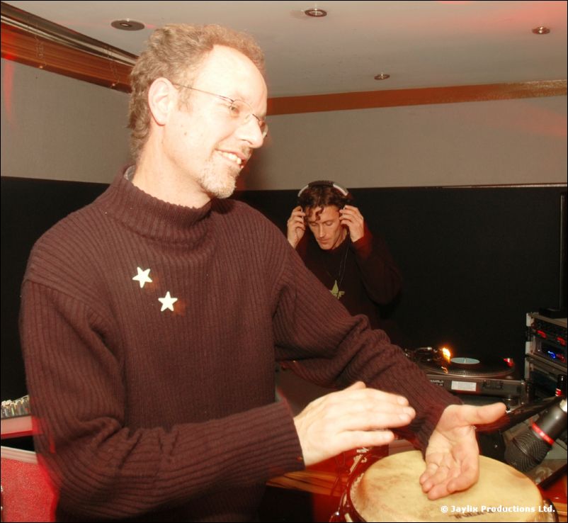 A man in a burgundy jumper with two stars plays a hand drum, with a DJ behind him