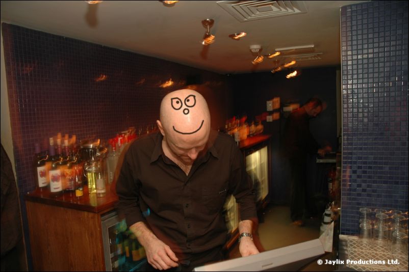 A man in a dark shirt with a smiley face drawn on his bald head stands behind a bar