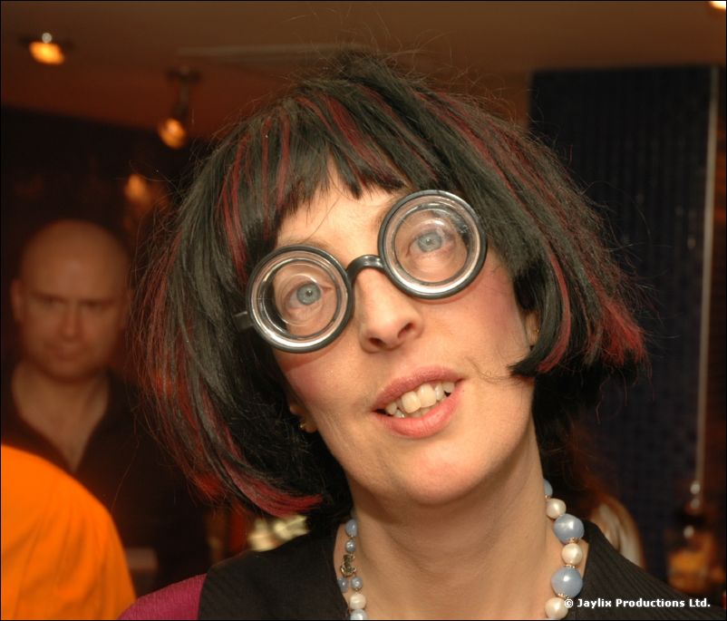 A woman with a dark bob and thick round glasses in a costume looks at the camera