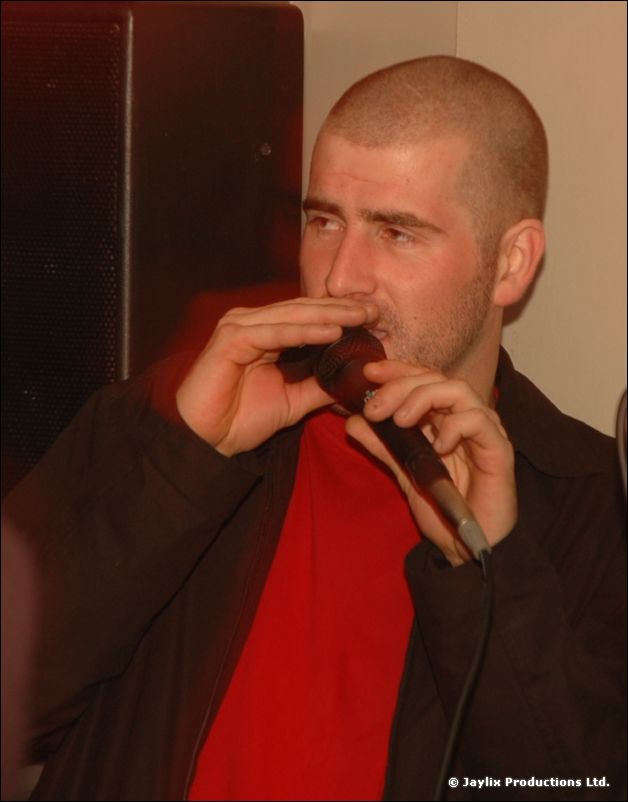 A man with a shaved head sings into a microphone, wearing a red top and dark jacket