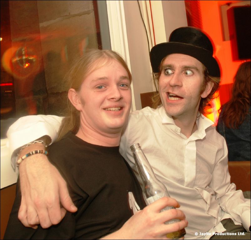 Two men hug and look at the camera, one in a black top hat and white shirt, the other in a black t-shirt