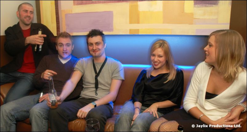 Five people sit on a sofa with drinks, looking to one side