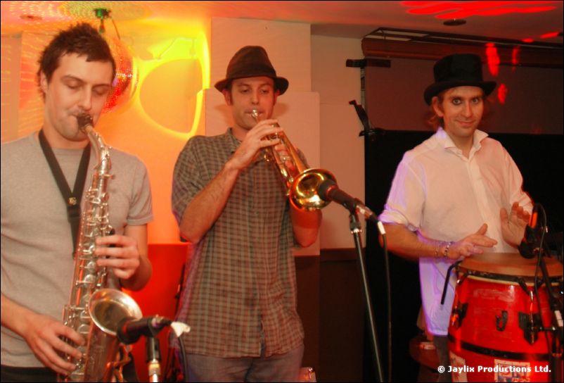 A saxophonist, a trumpeter and a man in a top hat playing congas perform together