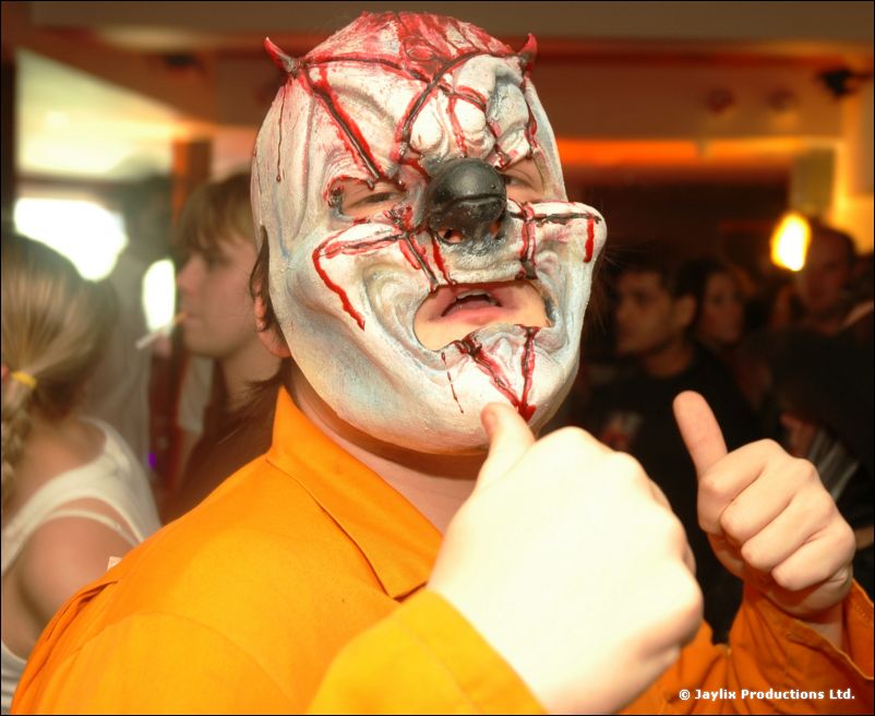 A person in a white bloodied horror mask and orange top gives two thumbs up