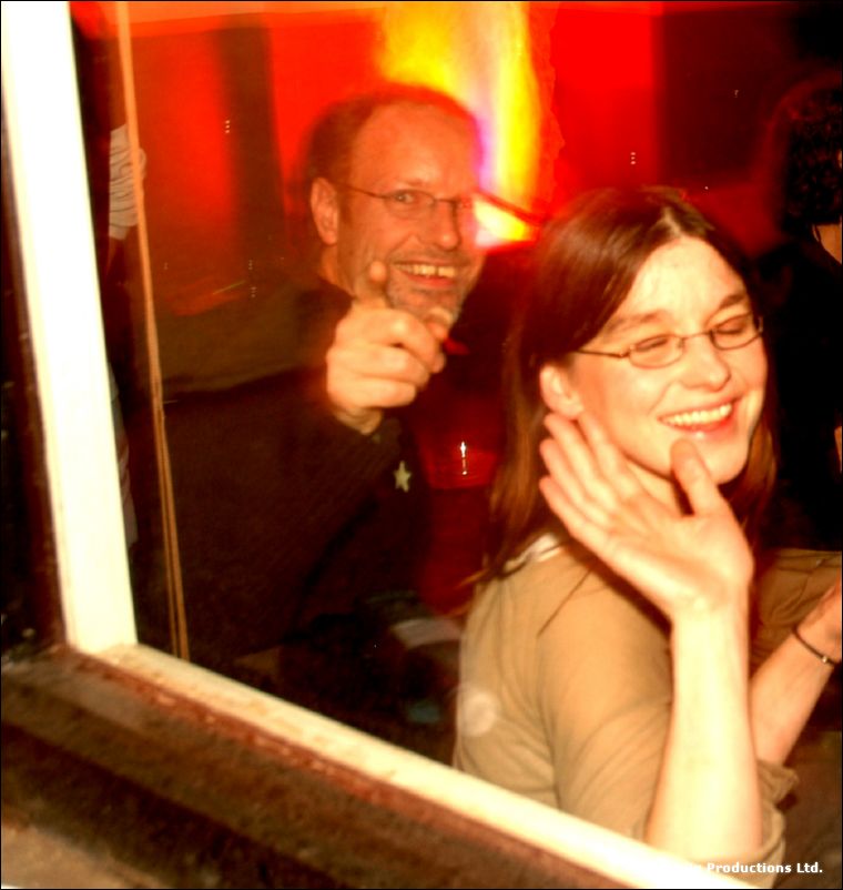 A man with glasses and a woman with glasses laugh behind a window pane