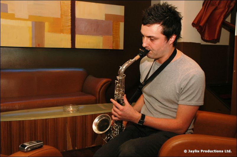 A man in a grey t-shirt plays a saxophone on a leather sofa