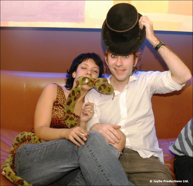A man in a top hat and a woman holding a toy snake sit together on a sofa