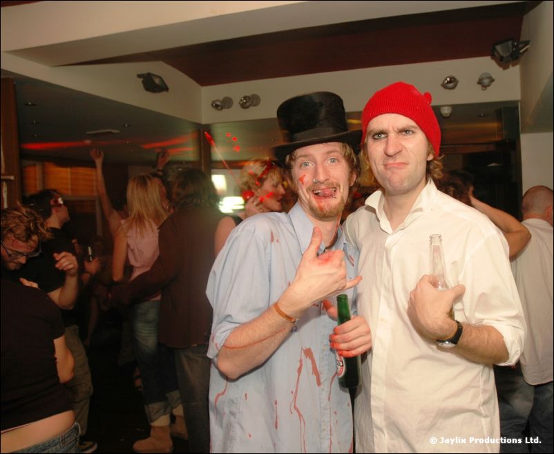 Two men pose together, one in a top hat and blood-spattered blue shirt, the other in a red beanie