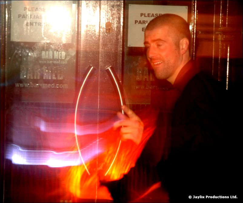 A smiling man in dark clothes swings a glowing fire prop in front of a Bar Med door