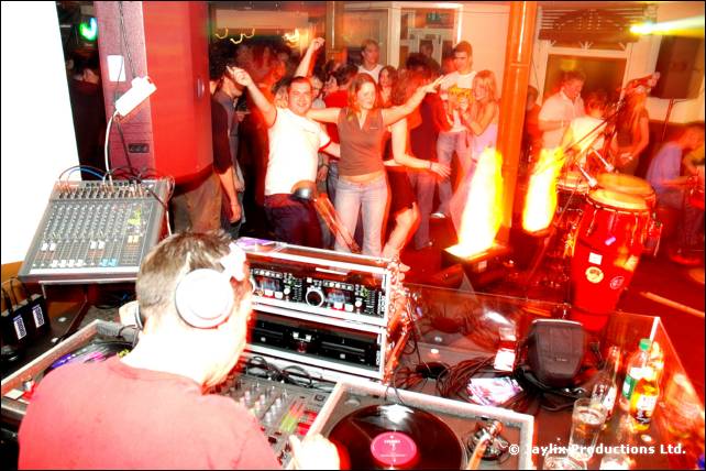 A DJ in a red top and headphones at mixing decks, with people dancing and congas behind