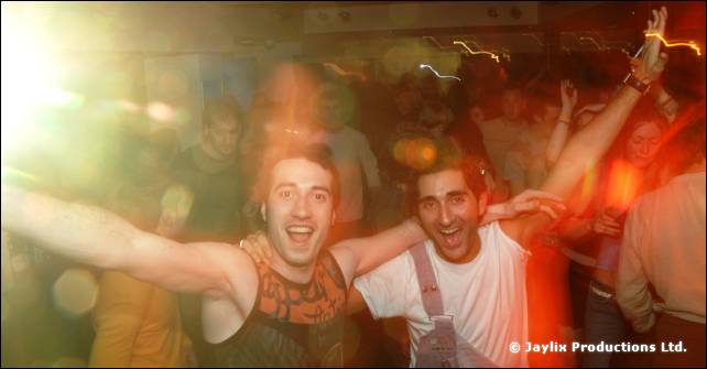 Two people with arms outstretched smile at the camera in a blurred, crowded room