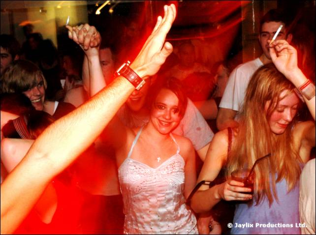 A smiling person with long hair holds a drink in front of a crowd, an arm raised in the foreground
