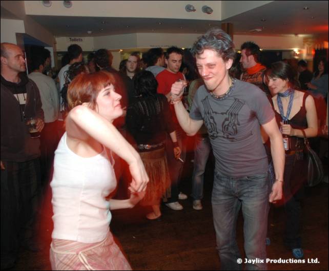 Two people dance in a busy bar, one in a white vest top and one in a grey t-shirt
