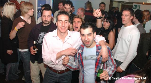 A crowded bar with two men in the foreground, one in a pink shirt and one in a checked shirt, smiling