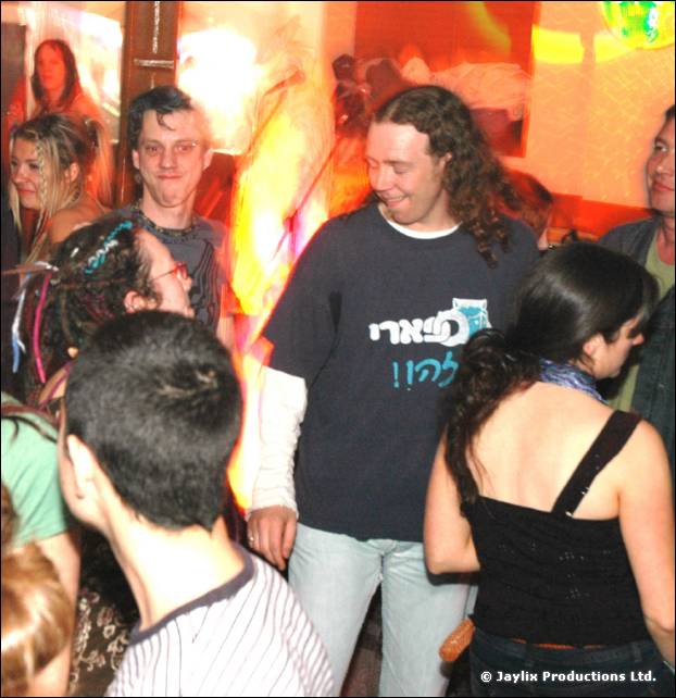 A person in a dark t-shirt with long curly hair smiles in a crowd beside a person with dark hair