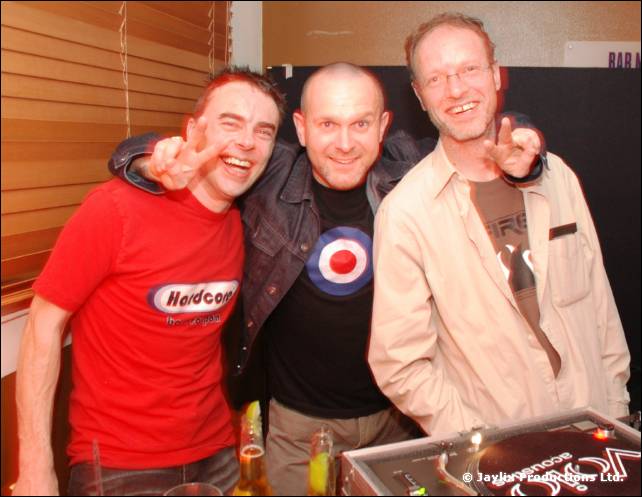 Three people with arms around each other, two making peace signs, stand behind DJ decks
