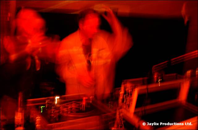 A heavily blurred scene with figures in red light behind DJ decks and equipment