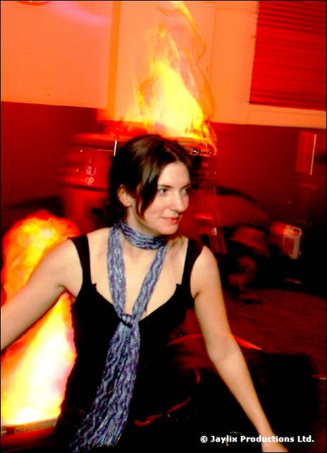 A person in a black top and blue scarf stands in front of a flame effect in a red lit room