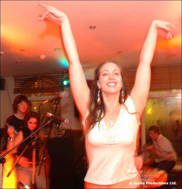 A person in a white top raises both arms and smiles, with seated people and microphones behind