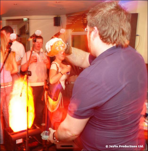 A with a bottle of orange drink in a crowded bar, a flame effect glowing behind