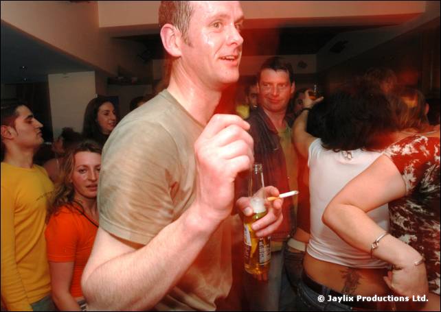 A person in a beige t-shirt holding a beer and cigarette stands in a crowd in a bar