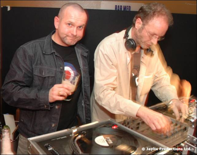 A person in a denim jacket holding a glass stands beside a DJ in a cream shirt at the decks