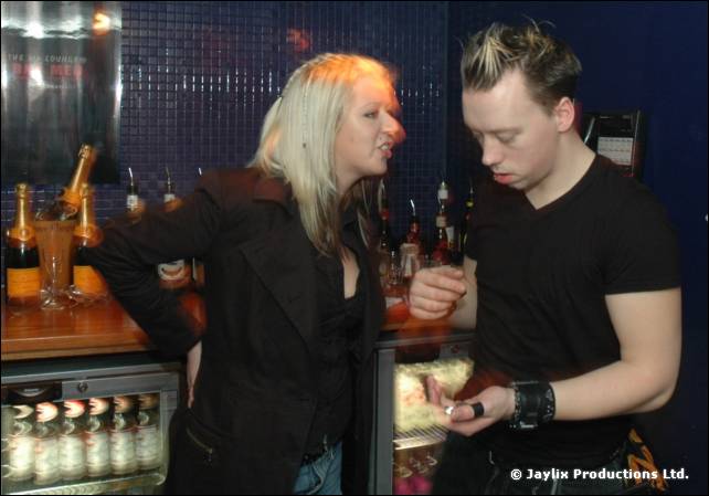 Two people stand at a bar with bottles behind, one with spiky hair in black holding a phone