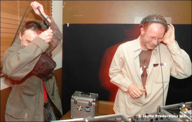 Two people at DJ decks, one in a cream shirt with headphones on, the other pulling a cable over his head