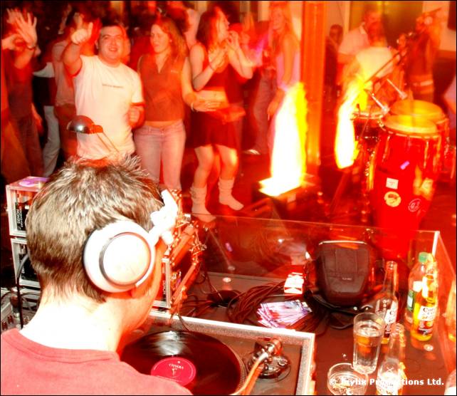 A DJ in a red top and headphones seen from behind, with a flame effect and congas beyond the decks