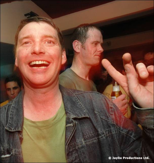 A laughing person in a denim jacket and green t-shirt, another person behind and a hand with pointing finger
