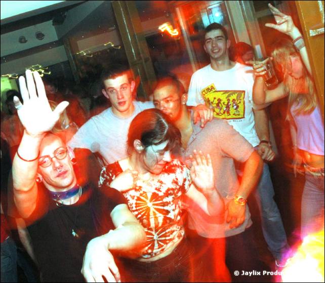 People dance in a crowded bar, one with arms raised and another in a yellow graphic t-shirt behind