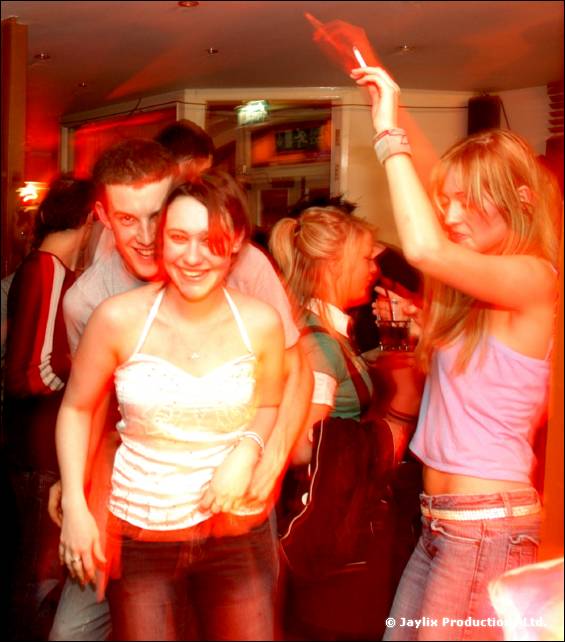 People dance closely in a red lit bar, one in a white top smiling, another in pink raising an arm with a cigarette