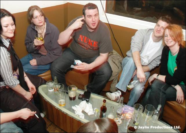 A group of people sit on a bench around a glass topped table with drinks, one holding a hand to their head