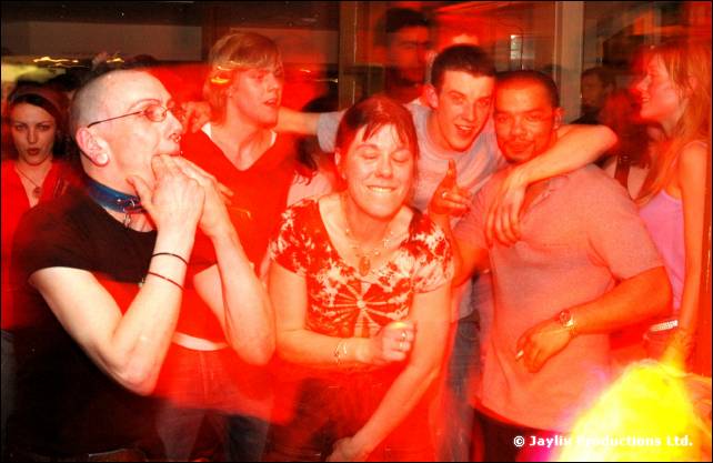 A crowd dances in red light, a person in glasses and black top in the foreground