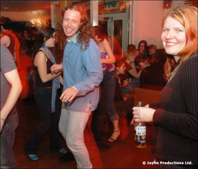 A man with long curly hair in a blue shirt dances beside a smiling woman holding a beer bottle