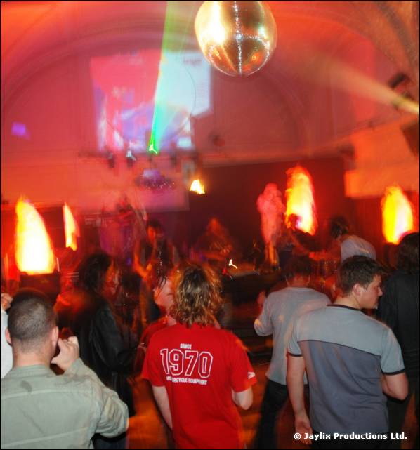 A crowd dances in a hall with a mirror ball, flame shaped lights and a projection, a man in a red 1970 top