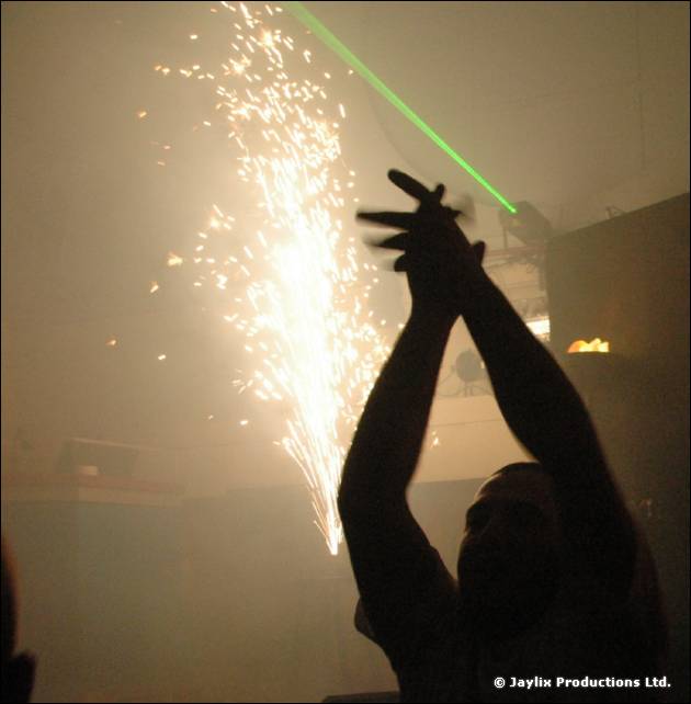 A silhouetted person claps above their head beside a spray of sparks and a green laser