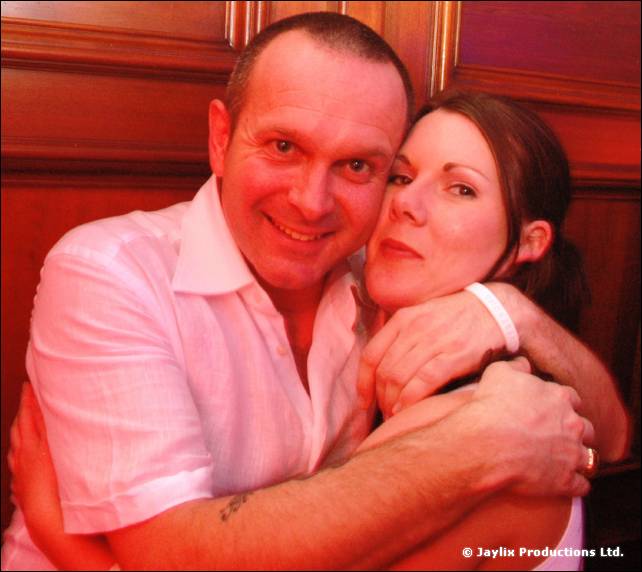 Smiling man in a white short-sleeved shirt hugs a woman with dark hair, in red light