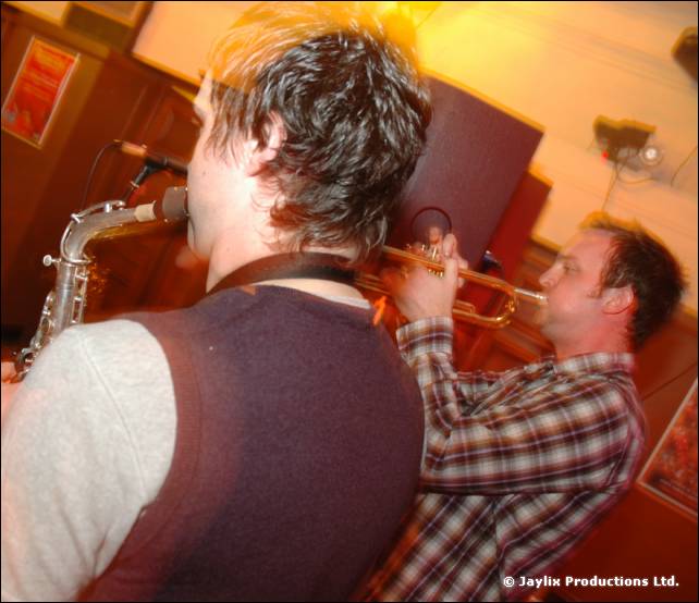 Two brass players, one on saxophone in a dark waistcoat and one on trumpet in a checked shirt