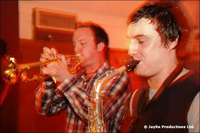 A man in a checked shirt plays trumpet beside a man in a dark waistcoat playing saxophone