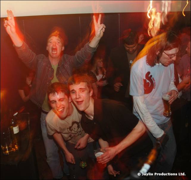 Crowd at a club night, one person making a peace sign, two smiling in front, and a person holding a pint