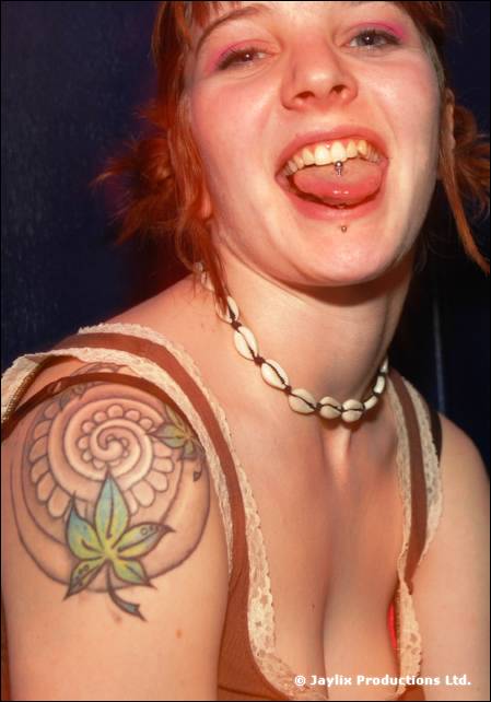 Smiling woman with red hair sticks out her tongue, showing a tongue stud and a leaf tattoo on her shoulder